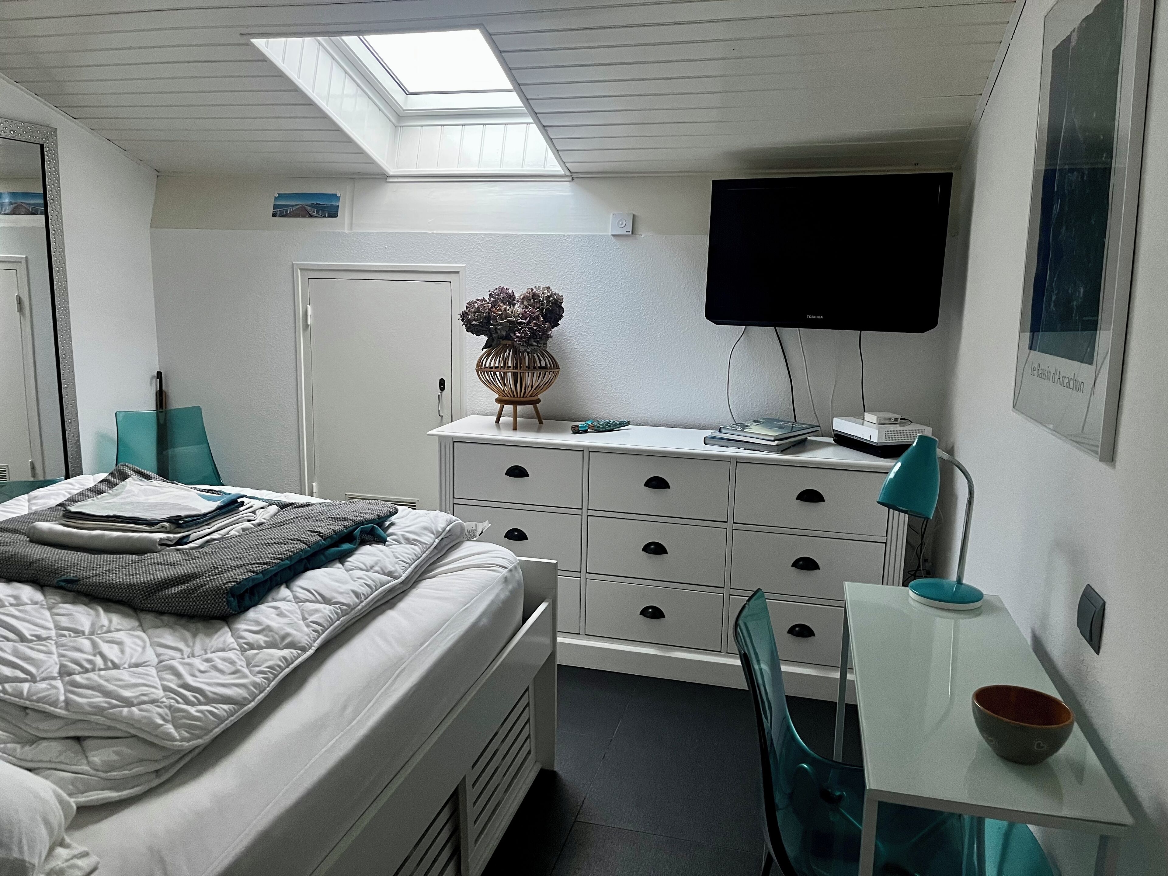 A bedroom with a bed, a desk, a chair, a television, and a skylight.