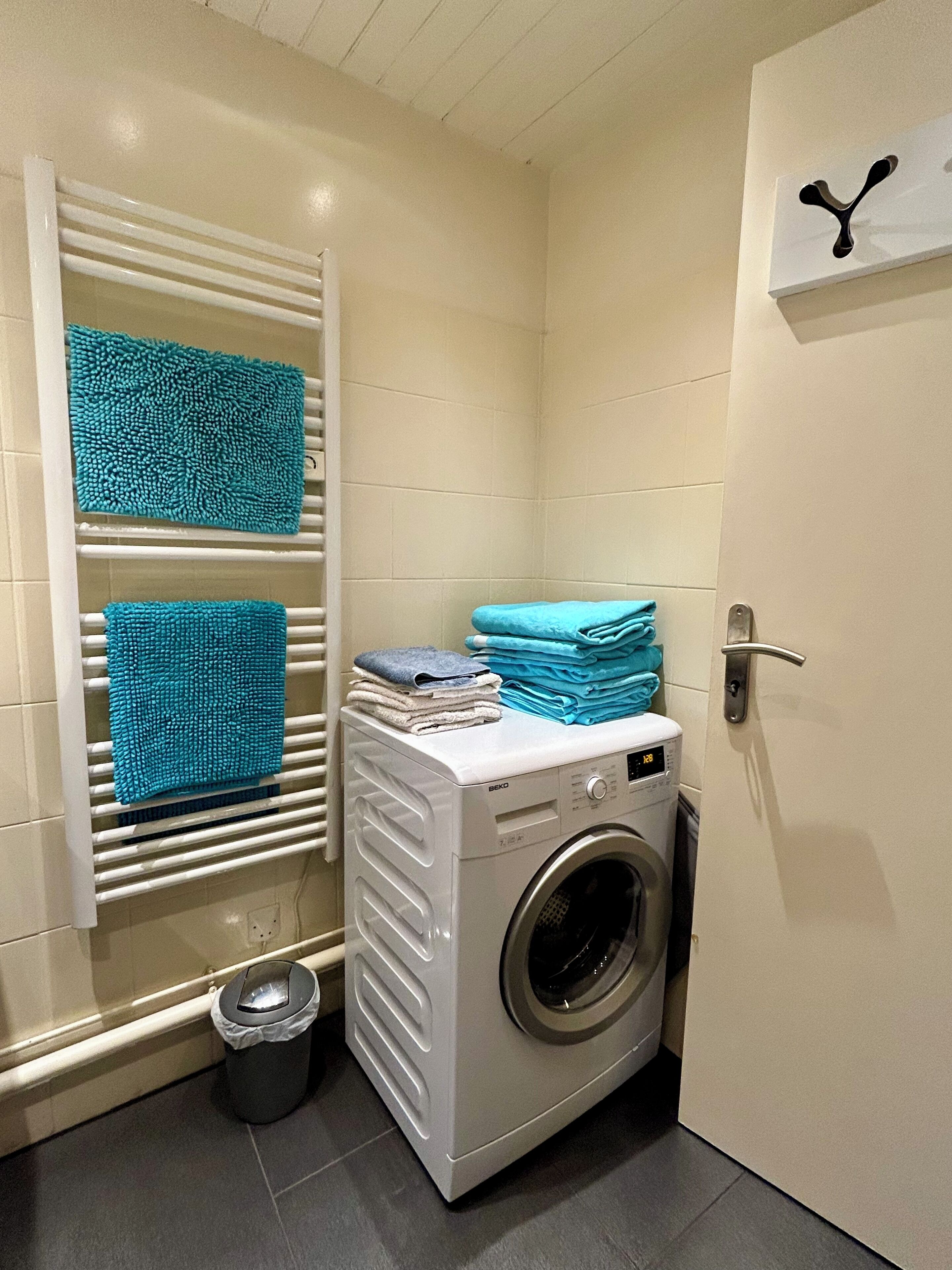 A compact laundry area with a washing machine, a drying rack with folded towels, and a small trash bin.