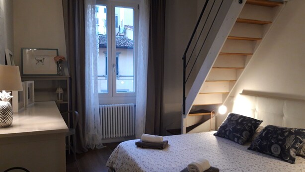 A bedroom with a bed, a desk, a radiator, a window with curtains, and a staircase.