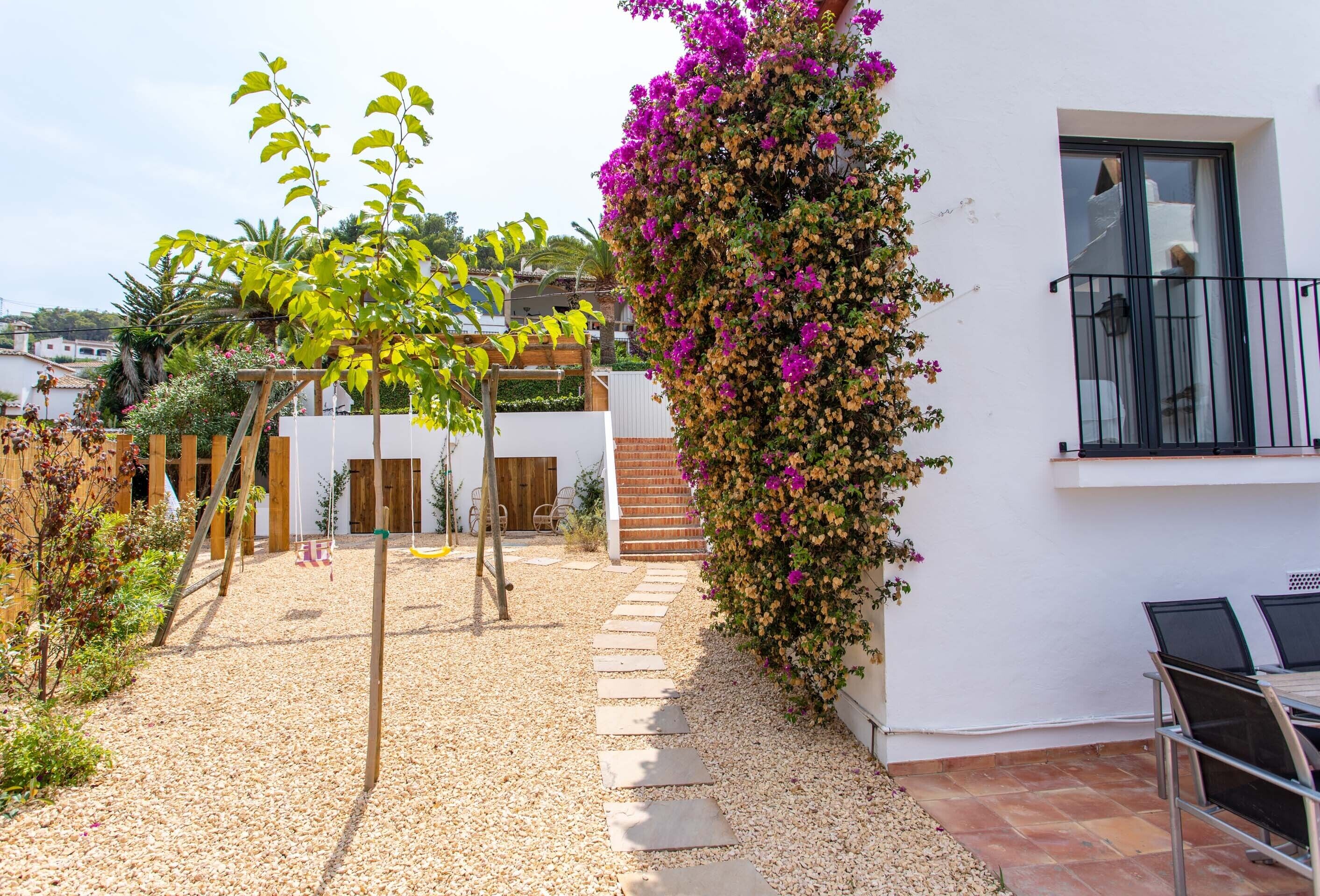 A courtyard with a gravel path, a wooden swing set, and a wall covered with flowering vines.