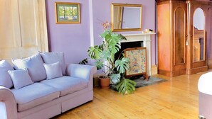 A living room with a light purple wall, a wooden floor, a light blue sofa, a large potted plant, a fireplace, and a wooden cabinet.