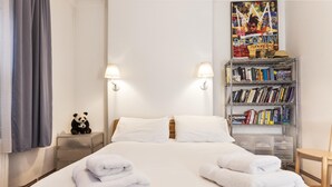 Ein Hotelzimmer mit einem ordentlich bezogenem Bett, zwei Nachttischlampen, einem Bücherregal mit Büchern, einer Panda-Figur und einem Poster an der Wand.