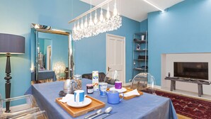 Ein Zimmer mit blauer Wand, einer Kristallleuchtre, einem Fernseher und einem Esstisch, der mit Tassen und einer Serviertablett gedeckt ist.
