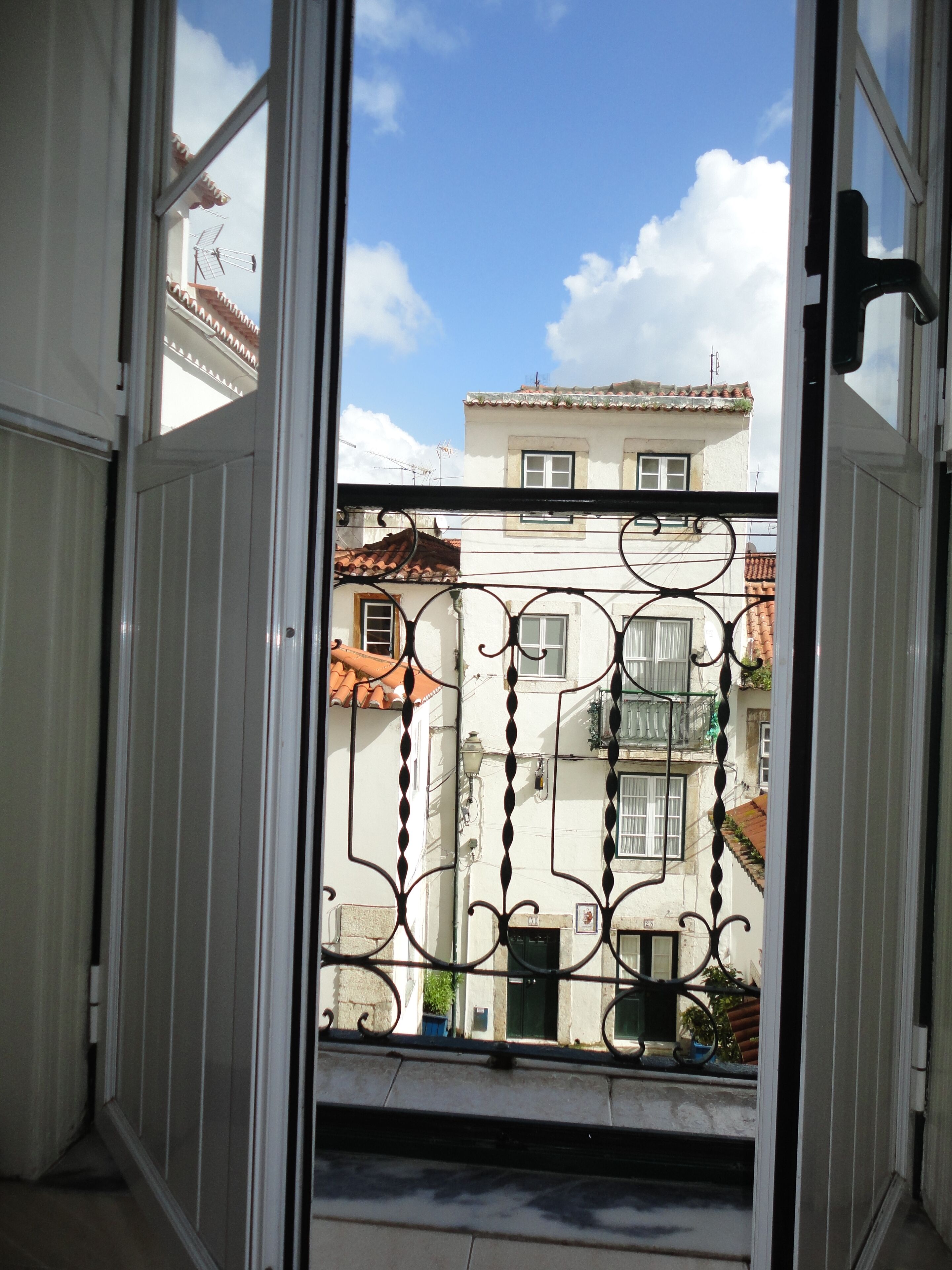 Uitzicht vanaf een balkon met een metalen hekwerk, uitkijkend op een straat met gebouwen en een heldere lucht.