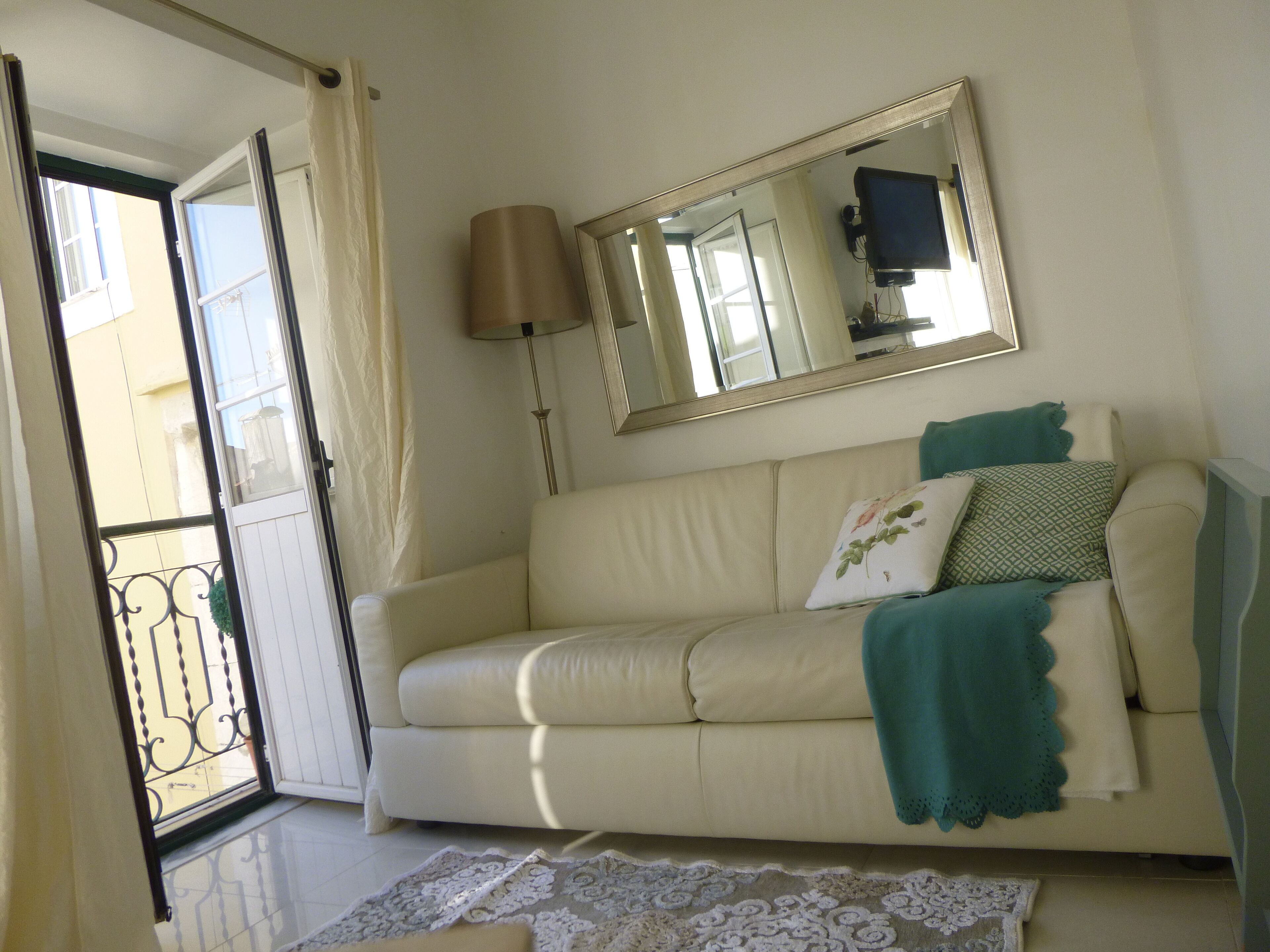 A living room with a beige sofa, a large mirror, a floor lamp, and a view through an open door.