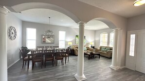 A spacious living room with a dining area, featuring high ceilings, large windows, and a modern chandelier.