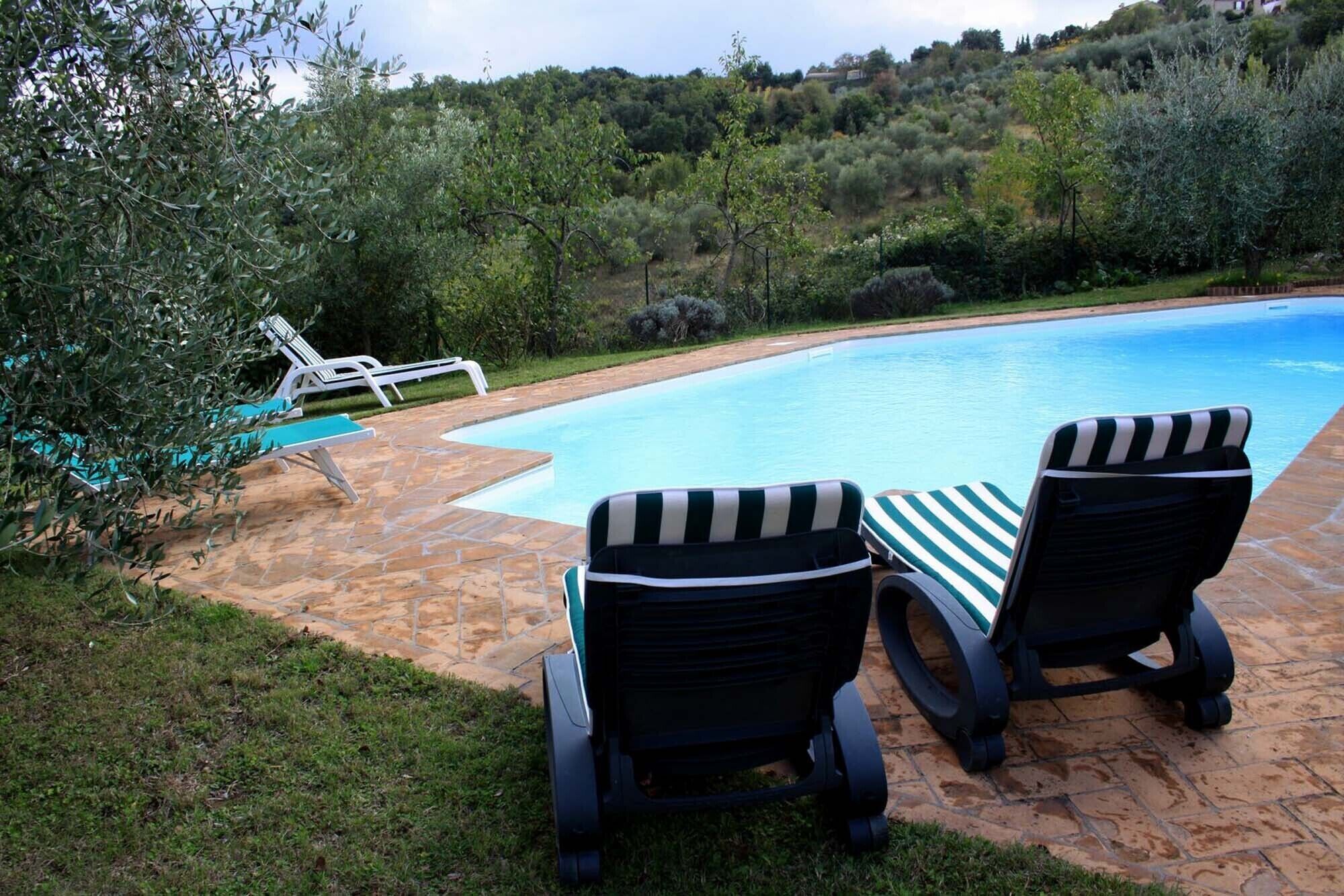 Une piscine bordée de deux chaises longues et d’une table, entourée d’une terrasse en pierre et d’une végétation luxuriante.
