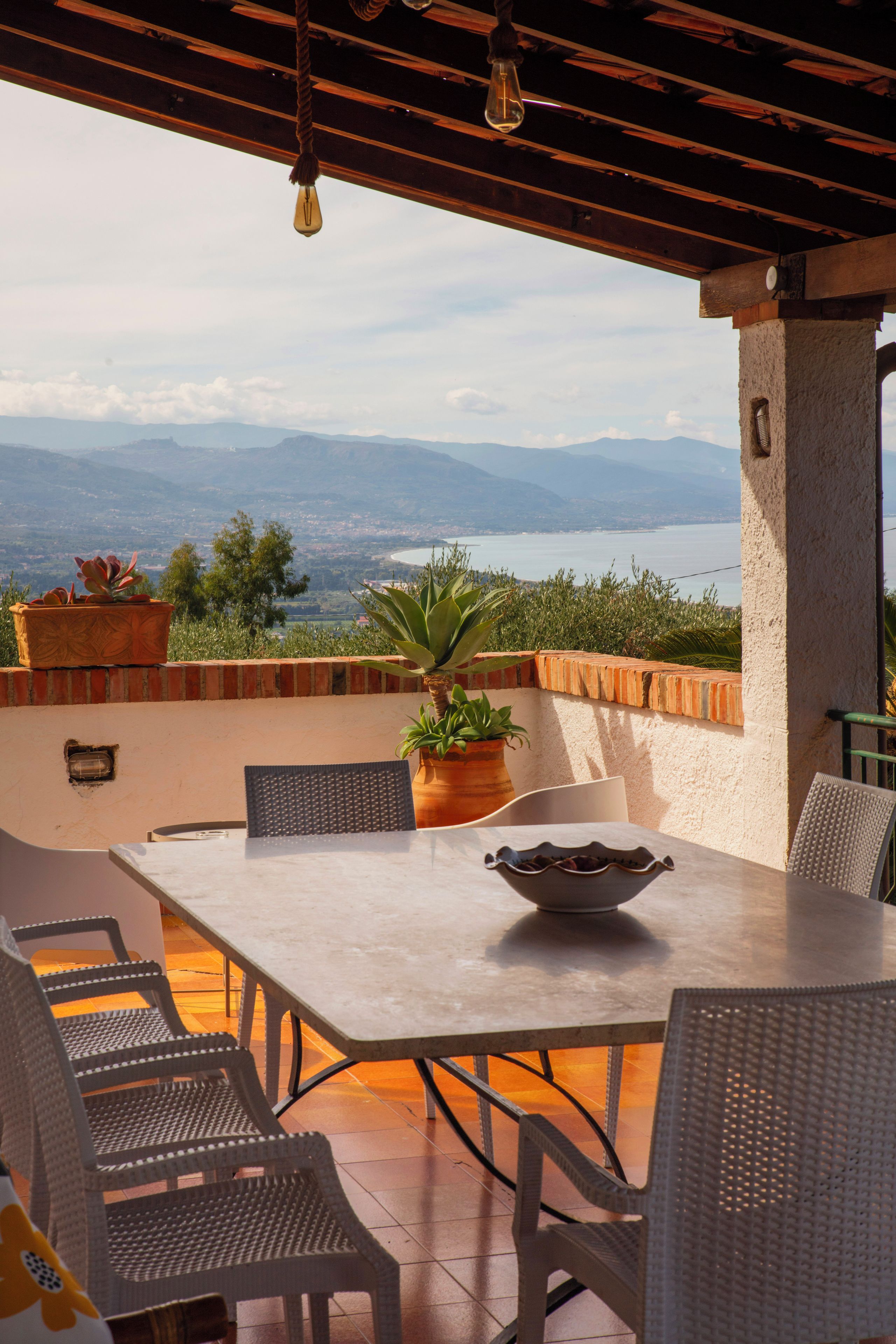 Une terrasse extérieure avec une table et des chaises, des plantes en pot et une vue sur un plan d’eau et des montagnes.