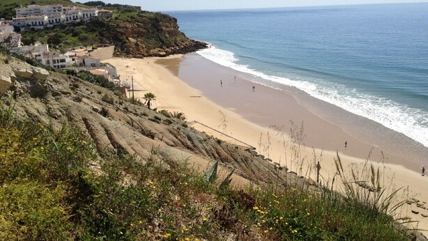 A coastal beach with a rocky cliff and buildings on the left.