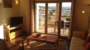 A living room with a television, a table, and a sofa. There is a balcony with a view of the countryside.