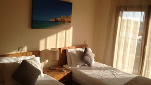 A hotel room with two beds, a nightstand with a lamp, a painting of a beach, and a window with curtains.