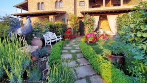 Ein Steinhaus mit einem Gartenweg, der zu einer weißen Bank und einer Hahn-Statue führt.
