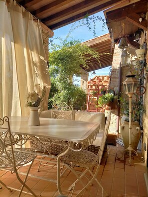 Une terrasse extérieure avec une table et des chaises, entourée de plantes en pot et d’un mur de pierre.