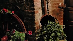 Ein rustikales Steinhaus mit einer Holztür, einem vergitterten Fenster und hängenden Laternen. Blumentöpfe mit roten Blüten und Grünpflanzen sind ebenfalls zu sehen.