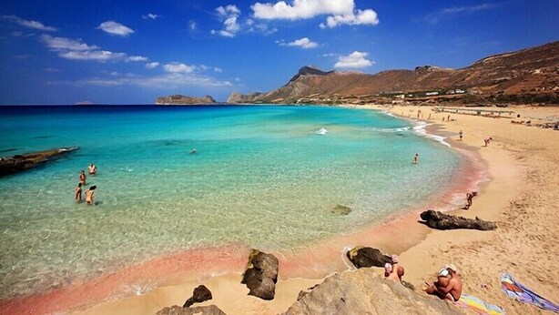 Ein Strand mit klarem türkisblauem Wasser, Felsformationen und einer bergigen Kulisse.