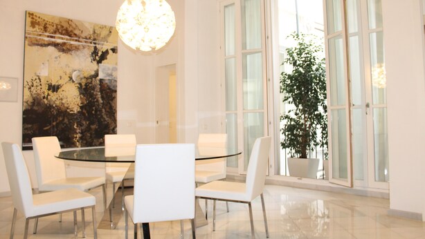 A modern dining area with a glass table surrounded by white chairs, a large abstract painting on the wall, and a potted plant near the window. - Unmatched Luxury next to the  Cathedral - Valencia (València)