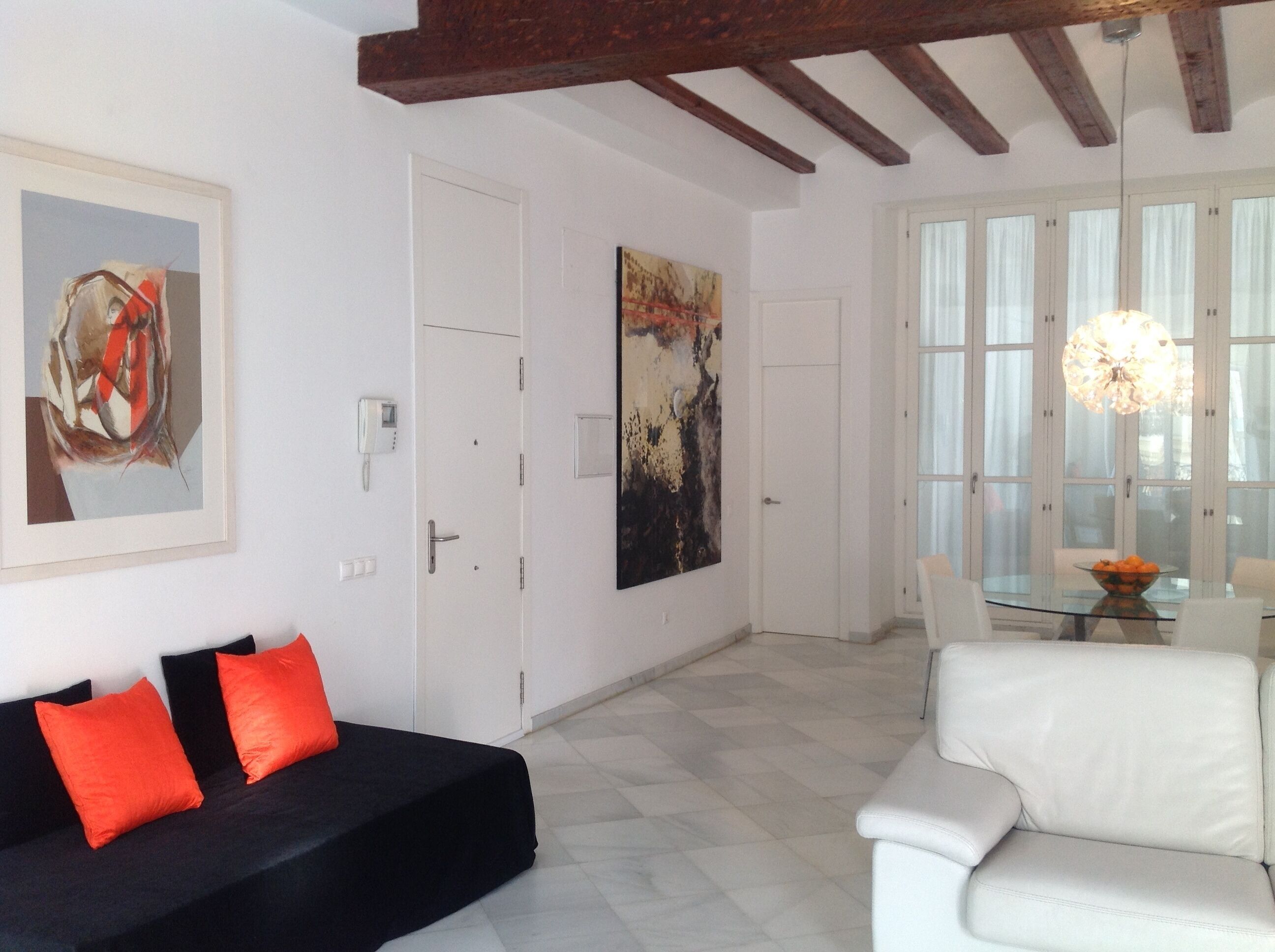 A living room with a black sofa, white armchair, and a glass table with a bowl of fruit. There are two paintings on the walls and a white door.