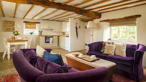 Una sala de estar acogedora con techo de madera, sofás morados, una mesa de comedor y una cocina al fondo.
