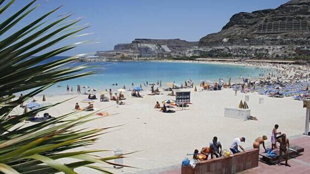 A beach with people sunbathing and swimming, a rocky cliff in the background, and a clear blue sky.