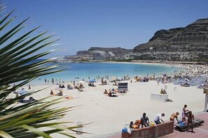 A beach with people sunbathing and swimming, a rocky cliff in the background, and a clear blue sky.