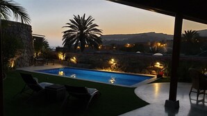 A swimming pool with a stone wall and palm trees, surrounded by lounge chairs and a stone wall.