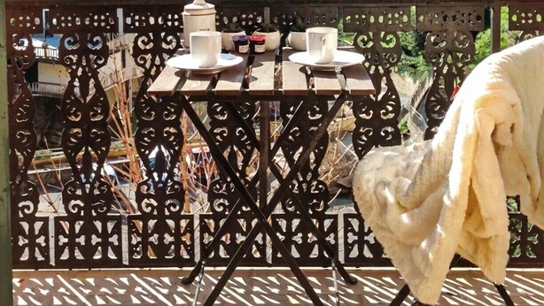 Un balcon avec une table dressée pour deux personnes, offrant une vue sur un village perché sur une colline, dont les bâtiments sont coiffés de toits en tuiles.