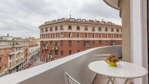 A balcony with a white table and a yellow flower vase, overlooking a row of red brick buildings with multiple windows and balconies. - Petrella (Milano)