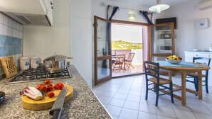 Una cocina con estufa a gas, tabla de cortar con verduras y comedor con mesa y sillas.