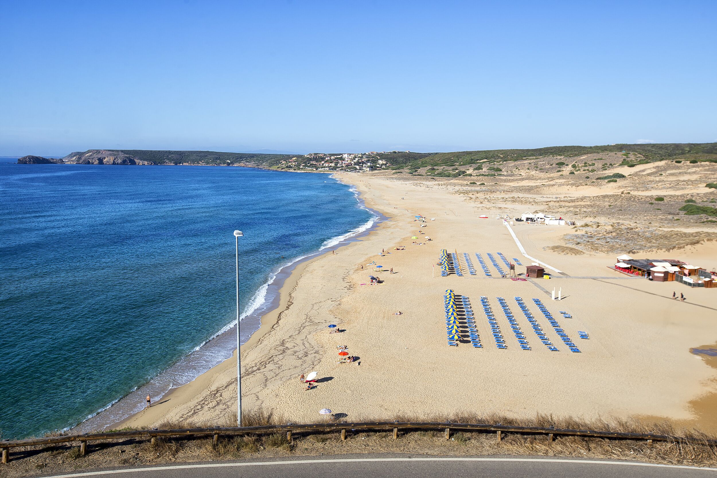 Une plage bordée de rangées de parasols bleus et de chaises longues, un rivage sablonneux et une mer d’un bleu limpide.