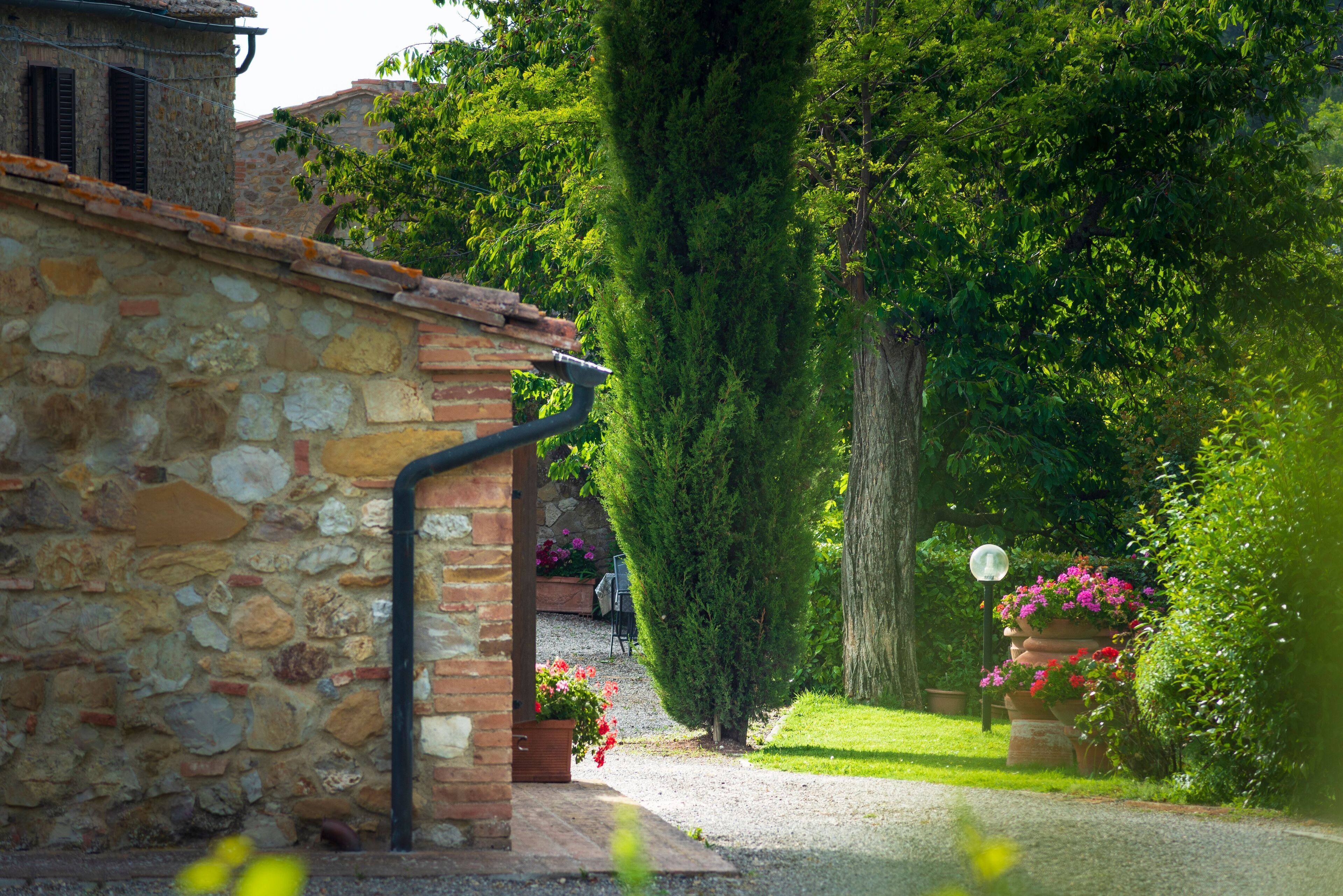 Un bâtiment en pierre avec un toit en briques, un chemin de gravier et une végétation luxuriante comprenant un grand arbre et des plantes à fleurs.