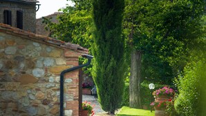 A stone building with a brick roof, a gravel path, and lush greenery including a tall tree and flowering plants.
