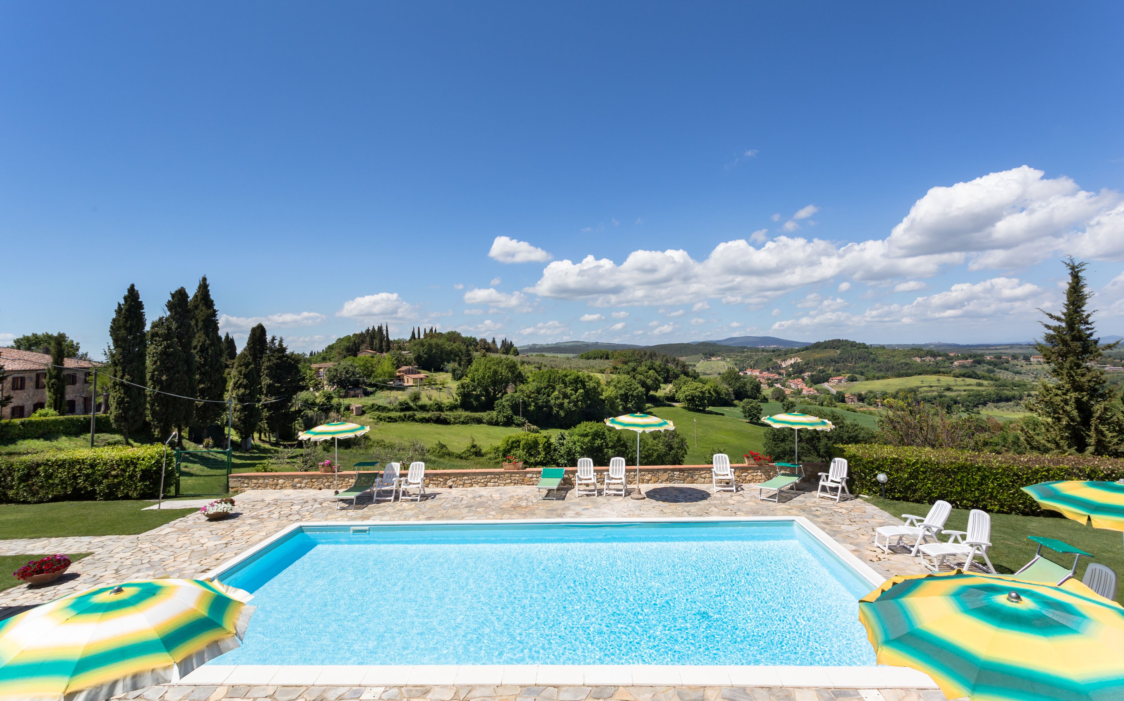 Une piscine rectangulaire bordée de pierres, entourée de chaises longues blanches et de parasols verts, offrant une vue panoramique sur un paysage pittoresque avec des collines et des arbres.