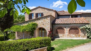 A stone building with a tiled roof, arched windows, and a garden with hedges.