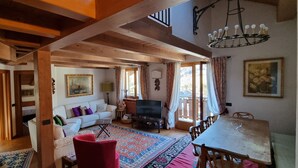 Een ruime woonkamer met een houten plafond, een witte bank, een rode fauteuil, een eettafel en een televisie.