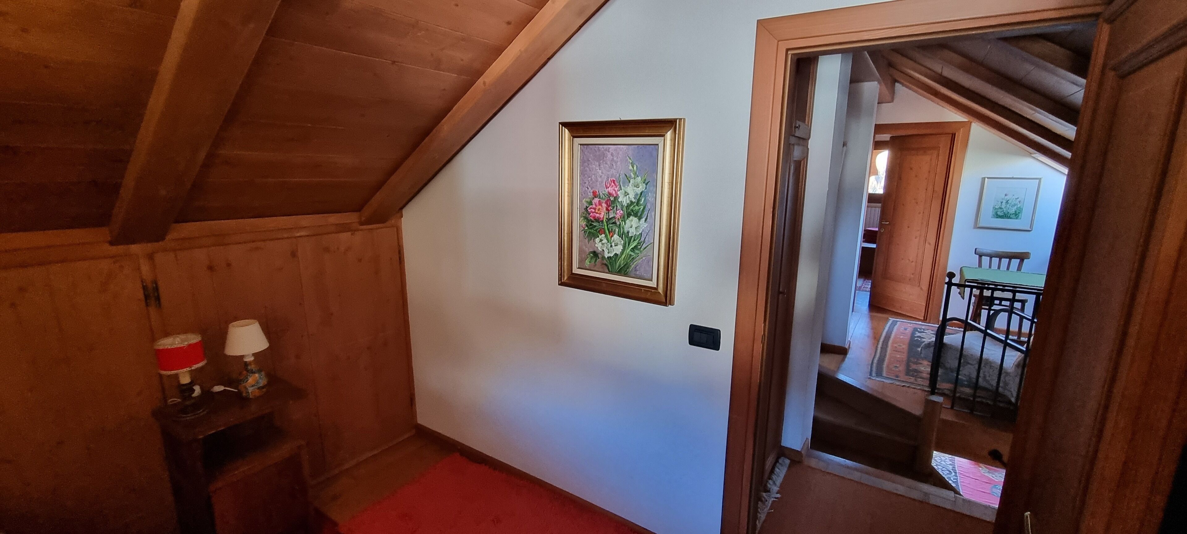 A hallway with wooden walls and a red carpet. There is a framed painting on the wall and a small table with two lamps. A staircase is visible in the background.