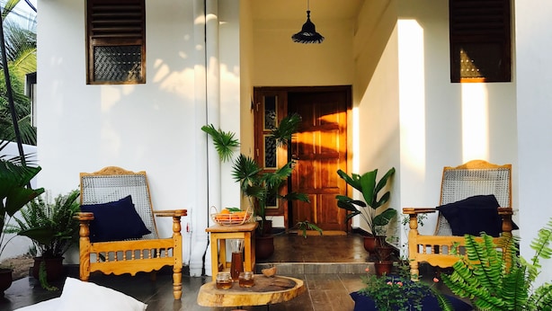 A traditional wooden chair with a cushion, a small table with a vase and jars, and potted plants in a well-lit hallway with a wooden door at the end. - Cozy Bay (Arugam Bay)
