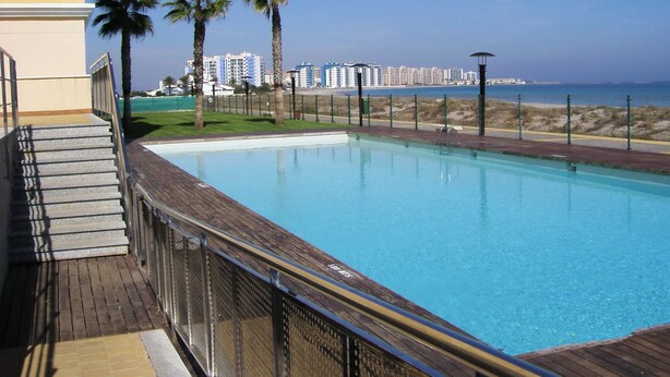 A swimming pool with a wooden deck and metal railing, surrounded by palm trees and a view of a beach and buildings in the background. - Beautiful, double BEACH FRONT Penthouse, Breathtaking View, Wi-Fi, SAT TV, Pool (San Javier)