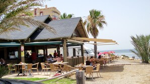 A beachside restaurant with outdoor seating, palm trees, and a view of the sea. - Beautiful, double BEACH FRONT Penthouse, Breathtaking View, Wi-Fi, SAT TV, Pool (San Javier)