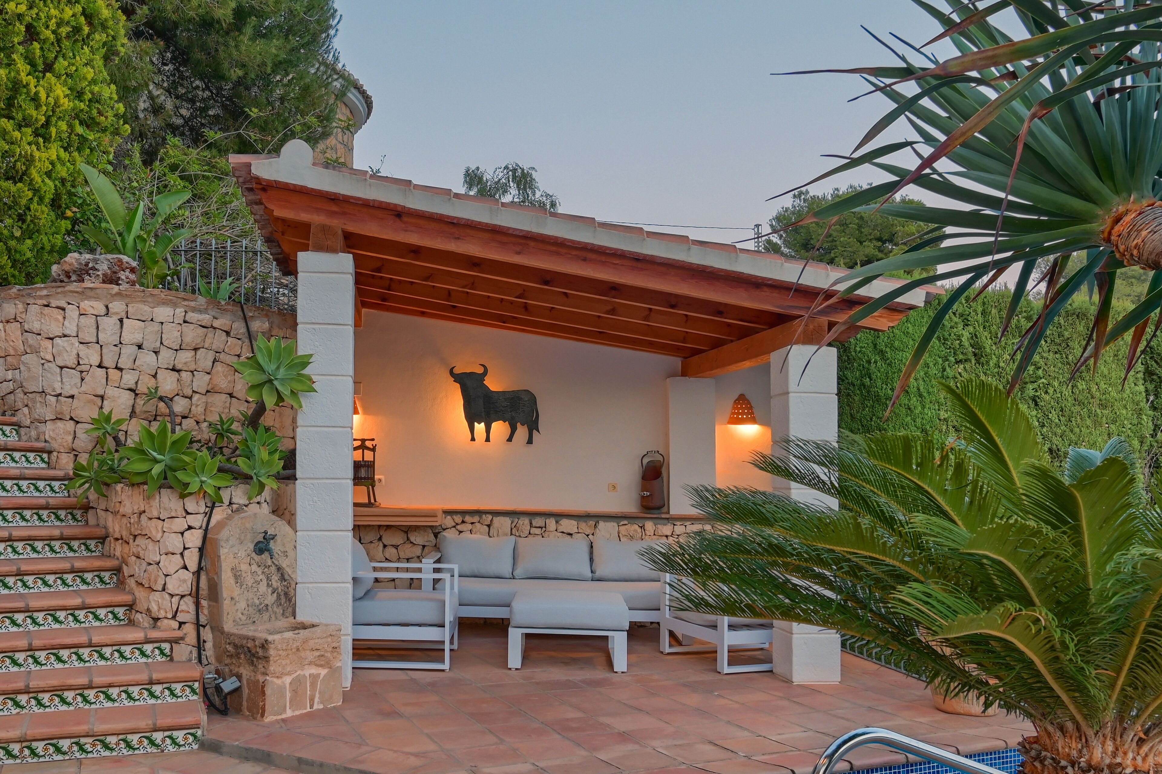 A covered outdoor seating area with a stone wall, a stone staircase, and a pool.