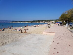 Een strand met mensen die zonnebaden en zwemmen, een heldere lucht en een verharde promenade.