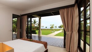 Un dormitori amb un llit, vista a un jardí i una platja, i una finestra gran amb cortines.