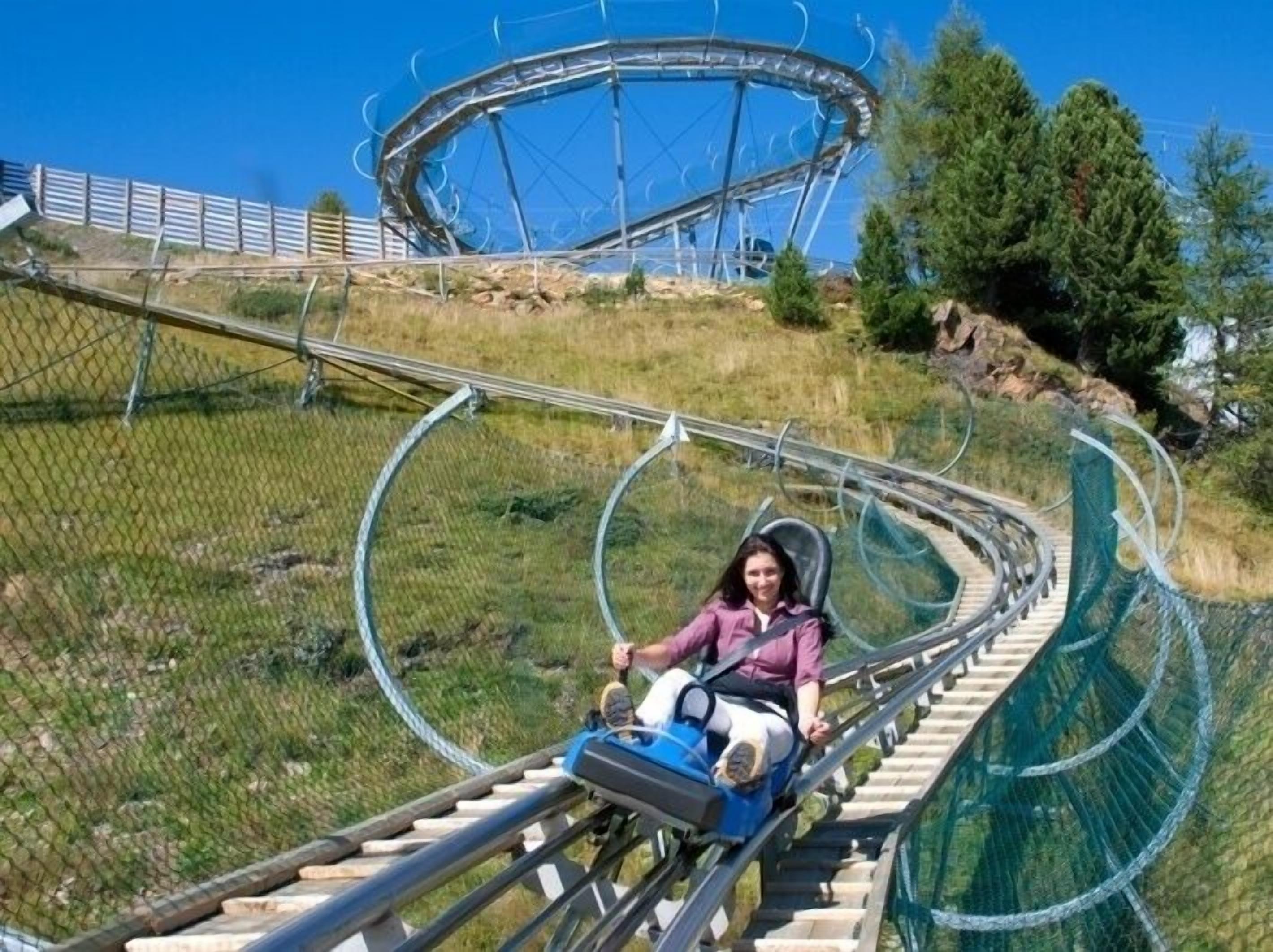 A person seated on a roller coaster track with a loop, surrounded by a fence and greenery.