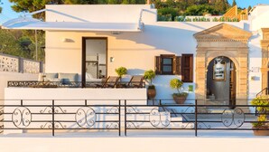 A building with a white facade, a balcony with sun loungers, a small entrance with a sign, and a view of a castle in the background. - Brand New!!! Lindos shore Superior Studio just above Lindos main beach !!! (Rhodes)