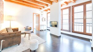 Un salon avec un canapé marron, une table ronde, un tapis blanc et un pouf moelleux. On y trouve un téléviseur posé sur un meuble blanc et une lampe fixée au mur. Le salon est doté de poutres en bois au plafond et de grandes fenêtres avec des cadres en bois.