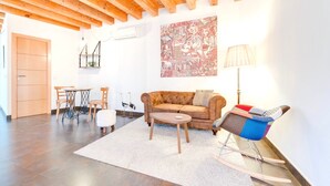 Un salon avec un canapé marron, une petite table ronde et un fauteuil à bascule. On y trouve un tapis blanc sur le sol, une porte en bois et un mur orné d’un grand tableau.