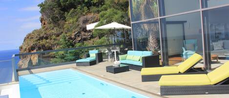 A modern outdoor pool area with lounge chairs and a table, overlooking a cliff and the sea.