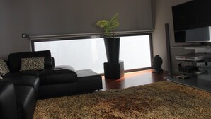 A modern living room with a large window, a black leather sofa, a potted plant, and a flat-screen TV. - Sea House Madeira - Atlantic Ocean (garajau)