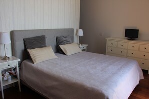 Un lit bien fait, recouvert d’une couvre-lit à motifs, avec deux oreillers, une lampe de chevet, un téléviseur posé sur une commode et une table de chevet sur laquelle se trouvent une lampe et un panier rempli d’objets divers.