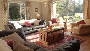 A living room with a black leather sofa, a wooden coffee table, and two armchairs. There is a window with curtains, a lamp, and a small table with flowers. - Fabulous location, luxury lodge for 8+2 in cots, hot tub, garden, garage, dogs   (Aviemore)