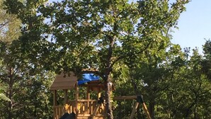 A playground with a wooden play structure, including a slide and swings, surrounded by trees and a stone wall. - Beautiful Provence House in the Luberon - Heated Swimming Pool & Large Gardens. (Roussillon)